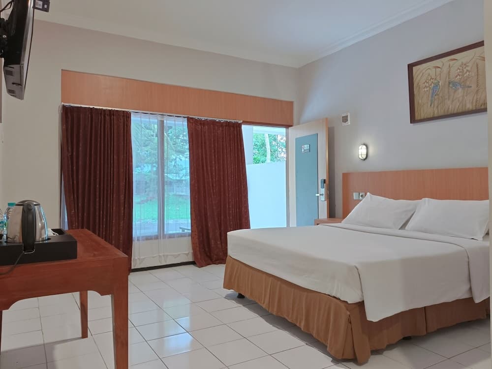 A cozy hotel room with a neatly made bed, wooden furniture, and a view of greenery through large windows, ideal for travelers seeking comfort and relaxation.