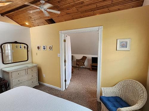 A cozy bedroom with warm yellow walls, wooden ceiling, and a wicker chair invites relaxation, perfect for travelers seeking a serene retreat in a vacation rental.