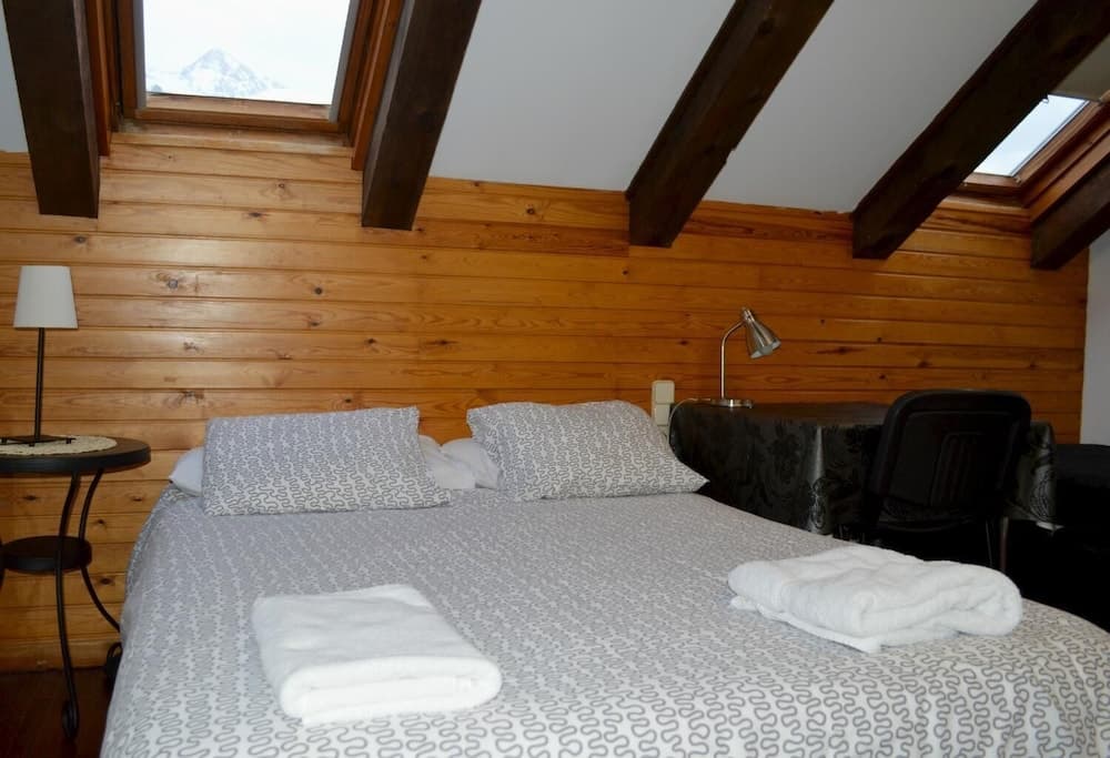 A cozy attic bedroom with wooden beams and walls, featuring a neatly made bed with patterned linens, white towels, and a scenic view of snowy mountains through the window, ideal for a serene mountain retreat.