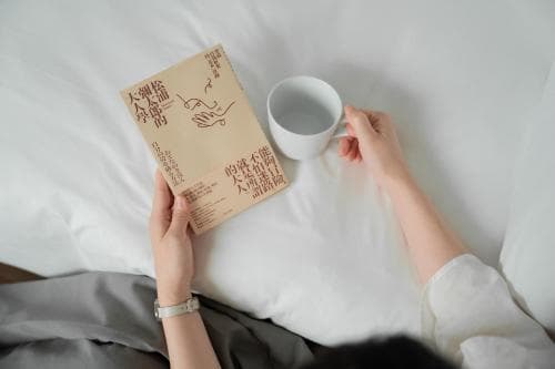 A cozy bedroom scene captures a relaxed moment with hands holding a book and a mug on white bedding, ideal for travel content promoting leisure and comfort.