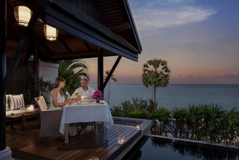 A couple enjoys an intimate dinner on a wooden deck overlooking the ocean at dusk, surrounded by lush tropical greenery and palm trees, creating a serene and romantic atmosphere ideal for a luxury beachfront getaway.
