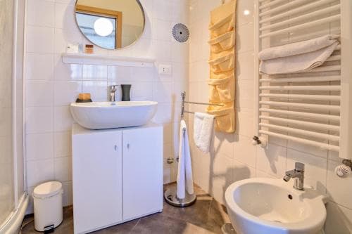 A cozy hotel bathroom features a white vanity sink, a toilet, neatly folded towels on a heated towel rack, and a round mirror, ideal for travelers seeking comfort and cleanliness during their stay.