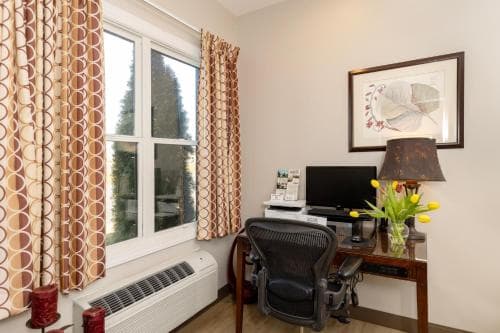 A cozy hotel room workspace features a wooden desk with a computer, ergonomic chair, vibrant yellow flowers, and patterned curtains, creating an inviting atmosphere ideal for business travelers seeking productivity.