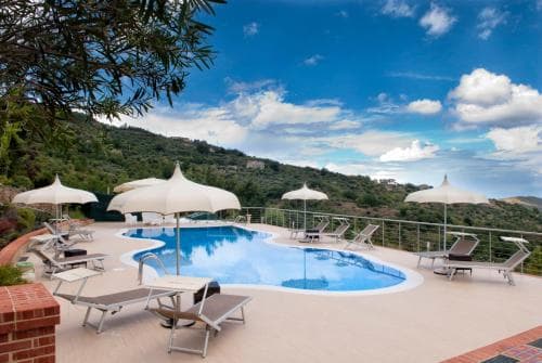 A serene outdoor pool area surrounded by lush hills under a vibrant blue sky, featuring lounge chairs, umbrellas, and a brick wall, ideal for a relaxing vacation retreat.