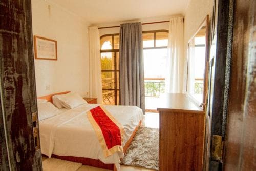 A cozy hotel room with a neatly made bed adorned with red accents, wooden furnishings, and large windows leading to a balcony, evokes a serene and inviting atmosphere ideal for travelers seeking relaxation.