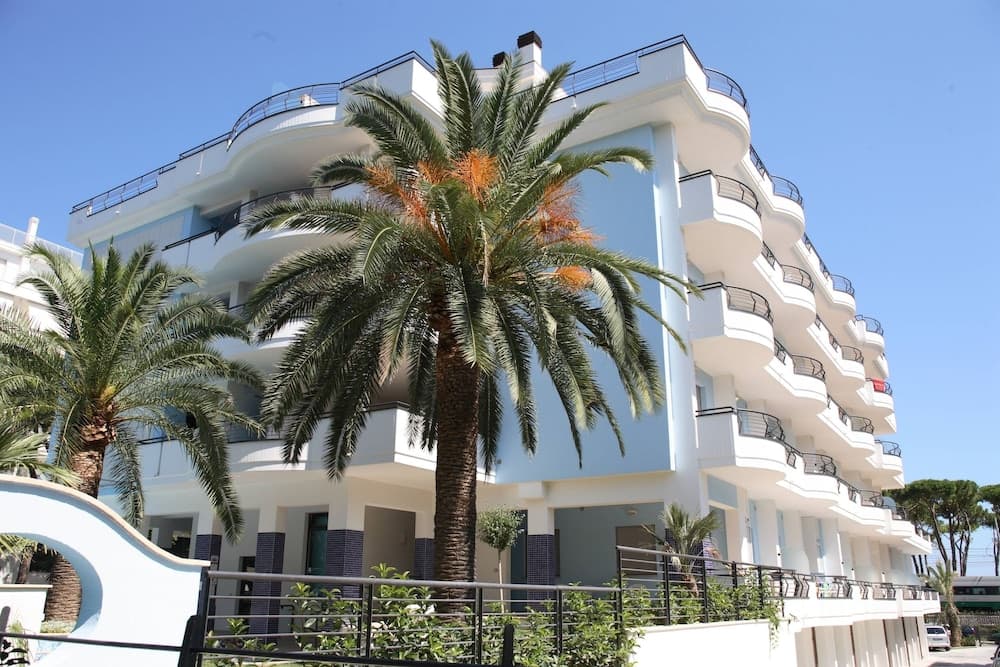 A serene coastal hotel scene features a modern white building with balconies, lush palm trees, and clear blue skies, ideal for travelers seeking a relaxing beachside getaway.