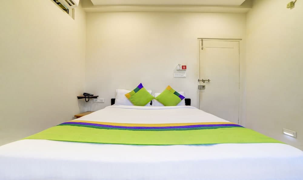 A cozy hotel room features a neatly made bed with vibrant green and purple accents, set against minimalist white walls, ideal for travelers seeking comfort and simplicity.