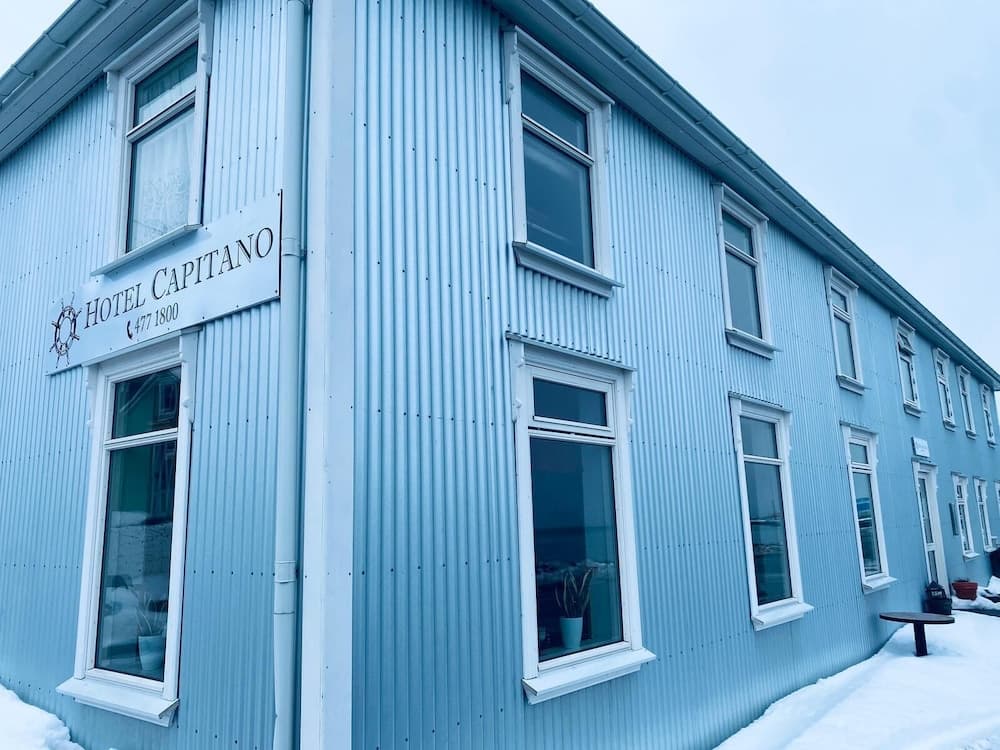 A serene winter scene showcases the light blue corrugated exterior of Hotel Capitano, featuring white-framed windows and a snow-covered pathway, ideal for travelers seeking cozy accommodations in a cold-weather destination.