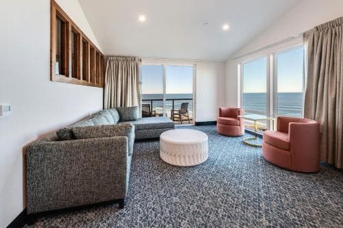 A cozy beachfront hotel room features a comfortable seating area with sofas and armchairs, large windows offering stunning ocean views, and a serene atmosphere ideal for relaxation and leisure travel.