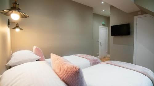 A cozy hotel room features two neatly made beds with pink accents, warm wall-mounted lamps, and a modern flat-screen TV, creating an inviting atmosphere ideal for travelers seeking comfort and relaxation.