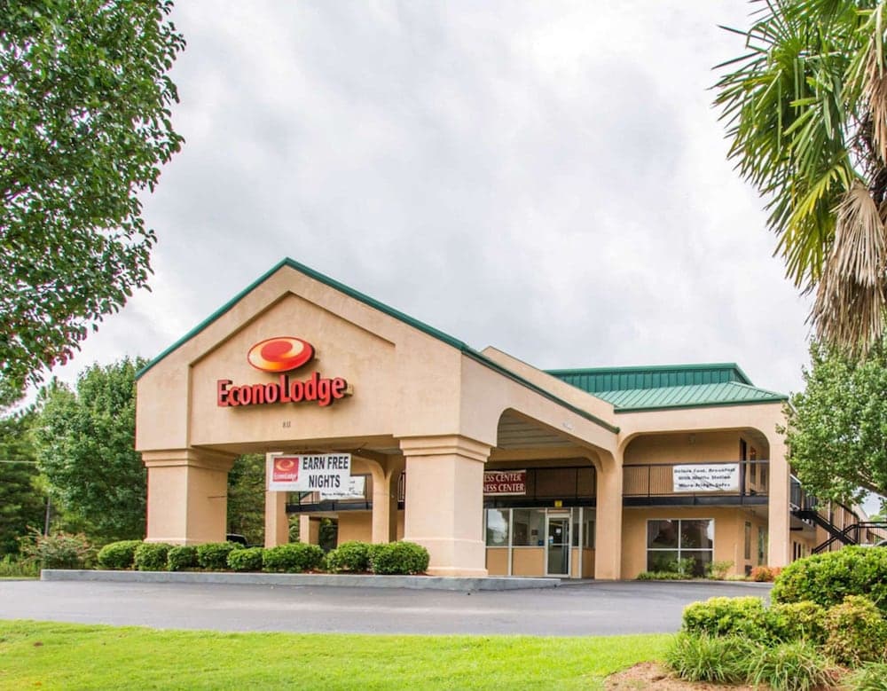 A welcoming Econo Lodge hotel entrance framed by lush greenery under an overcast sky, featuring promotional signs and a business center, ideal for travelers seeking budget-friendly accommodations.