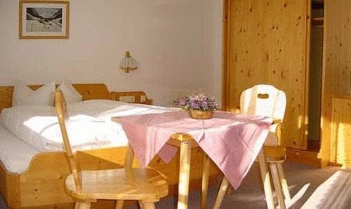 A cozy wooden-themed hotel room features a neatly made bed, a small dining table set with a pink tablecloth and flowers, evoking a warm and inviting atmosphere ideal for travelers seeking comfort and simplicity.