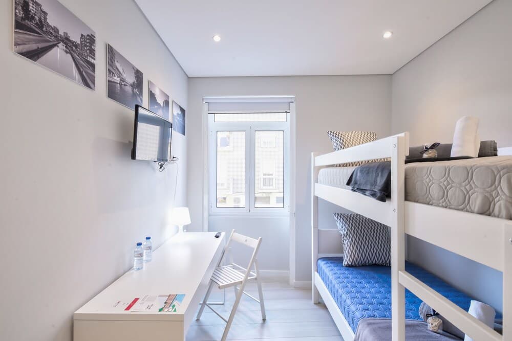 A cozy hostel room features bunk beds with blue and gray bedding, a sleek white desk with a chair, a mounted TV, and framed cityscape photos, creating a modern and inviting atmosphere ideal for budget travelers seeking comfort.