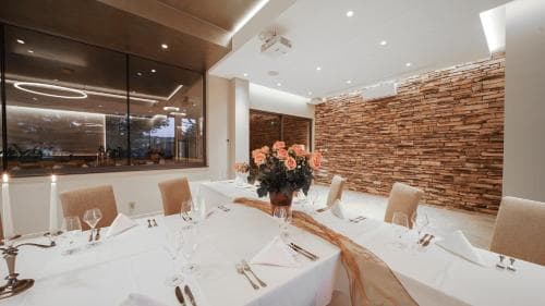 A modern dining room with a rustic brick wall and sleek glass windows sets an elegant mood, featuring a long table adorned with floral arrangements and fine dining ware, ideal for corporate events or upscale gatherings.
