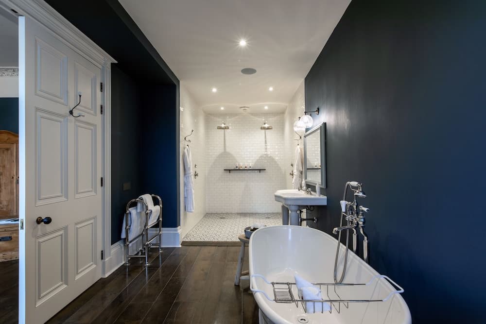 A luxurious bathroom with dark blue walls, a freestanding bathtub, white tiled shower area, and wooden flooring creates an elegant and serene atmosphere ideal for high-end accommodation or spa retreats.
