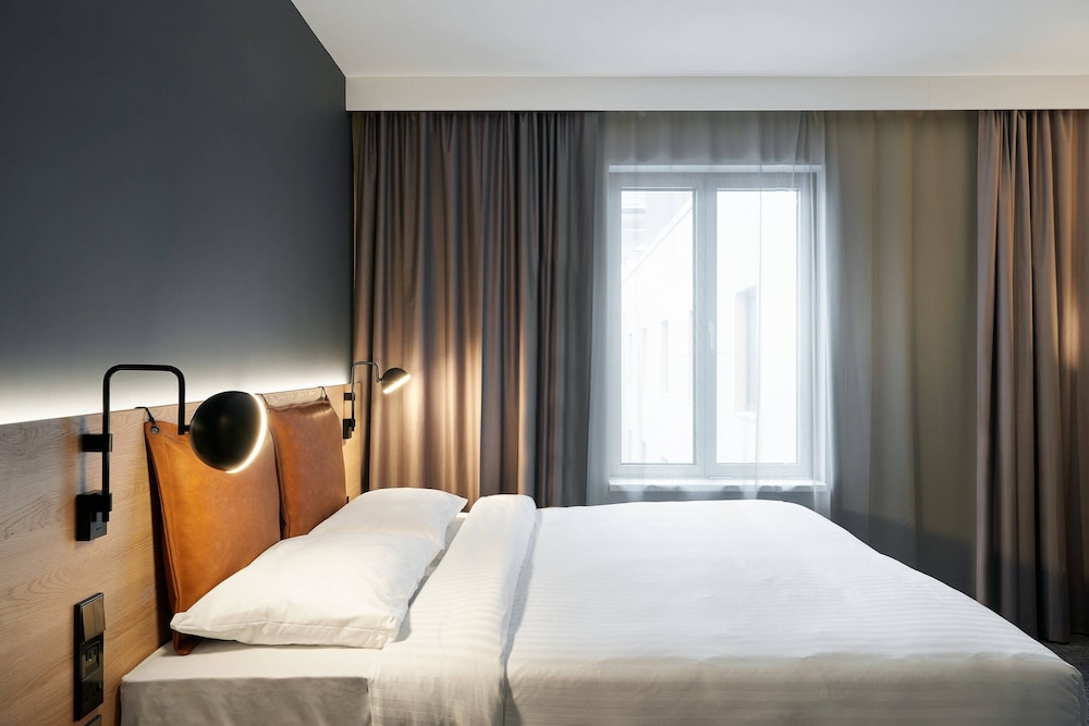 A serene hotel room features a neatly made bed with white linens, modern wall-mounted lamps, and a large window with sheer curtains, ideal for travelers seeking comfort and relaxation.
