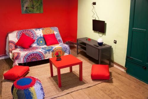 A cozy living room with vibrant red walls, colorful patterned furniture, and wooden floors creates a warm and inviting atmosphere, perfect for travelers seeking a comfortable and artistic accommodation.