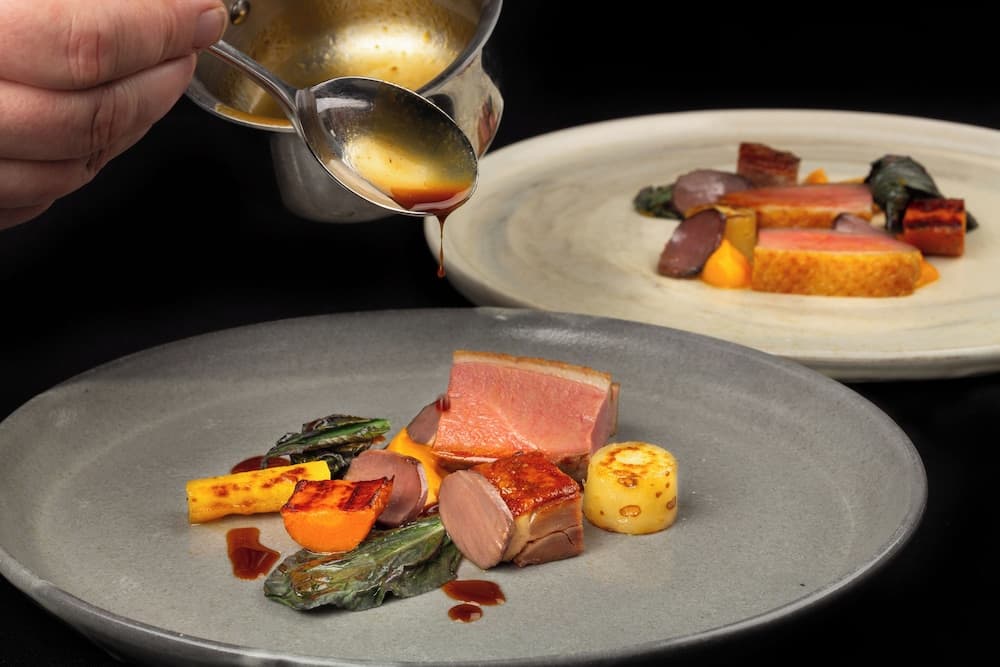 A chef artfully pours sauce over a gourmet dish featuring seared duck breast, roasted vegetables, and a potato galette on a sleek plate, perfect for culinary enthusiasts exploring fine dining experiences.