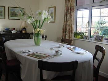 A cozy dining room bathed in natural light features a set table with white linens, a vase of elegant lilies, and framed artwork, creating an inviting atmosphere perfect for a serene breakfast or intimate gathering.