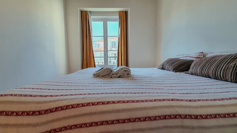 A cozy hostel room in Lisbon features a neatly made bed with striped bedding, folded towels, and a window offering a serene view of traditional rooftops, ideal for travelers seeking comfort and charm.