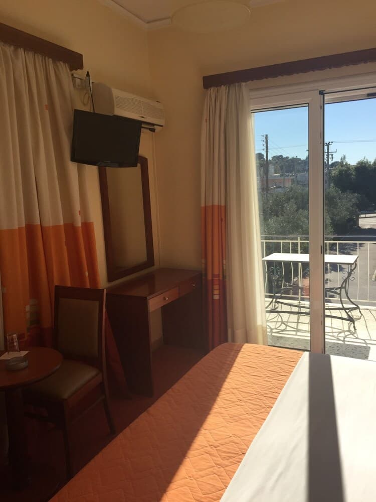 A cozy hotel room with warm tones, a wooden desk, flat-screen TV, and a sunny balcony view invites travelers seeking comfort and relaxation.