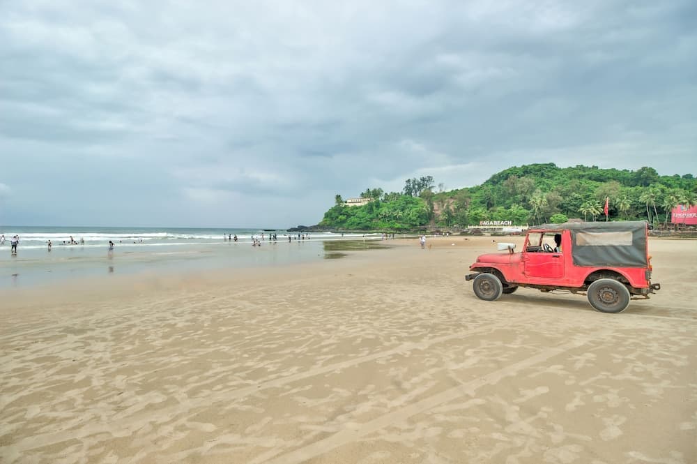 A serene beach scene under cloudy skies features a red jeep parked on the sand near gentle waves, with lush green hills in the background, ideal for adventure travelers seeking coastal exploration.
