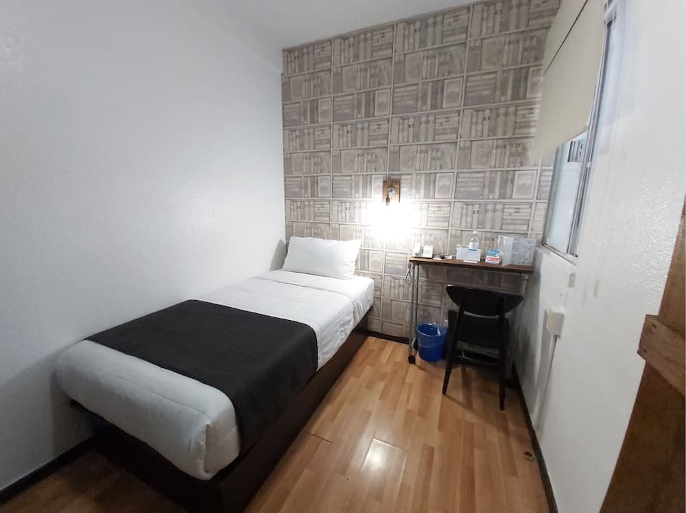 A cozy, minimalist hotel room features a neatly made bed against a bookshelf-patterned wall, a small desk with amenities, and warm wooden flooring, ideal for budget travelers seeking comfort and simplicity.