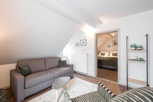 A cozy attic apartment features a modern living space with a gray sofa, wooden flooring, and an open view into a bedroom, ideal for travelers seeking a stylish and comfortable urban retreat.