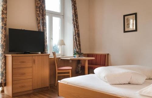 A cozy hotel room features a neatly made bed, wooden furniture, a flat-screen TV, and soft natural light filtering through patterned curtains, ideal for travelers seeking comfort and relaxation.