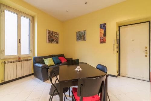 A cozy apartment living room with yellow walls, a leather sofa adorned with colorful pillows, a dining table set for two, and framed artwork, ideal for travelers seeking a comfortable urban retreat.