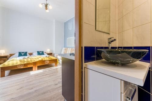 A cozy bedroom with a wooden bed adorned in warm yellow bedding is partially visible through an open doorway, adjacent to a modern bathroom featuring a sleek black vessel sink and blue-tiled walls, ideal for travelers seeking comfort and style in their accommodations.