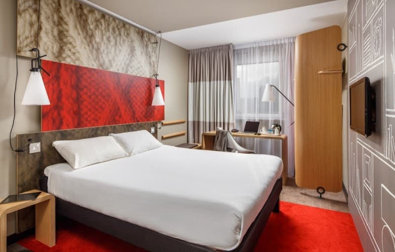 A cozy hotel room at the ibis Amsterdam Centre features a neatly made bed, vibrant red accents, modern furnishings, and a bright window view, ideal for travelers seeking comfort and style in the heart of Amsterdam.