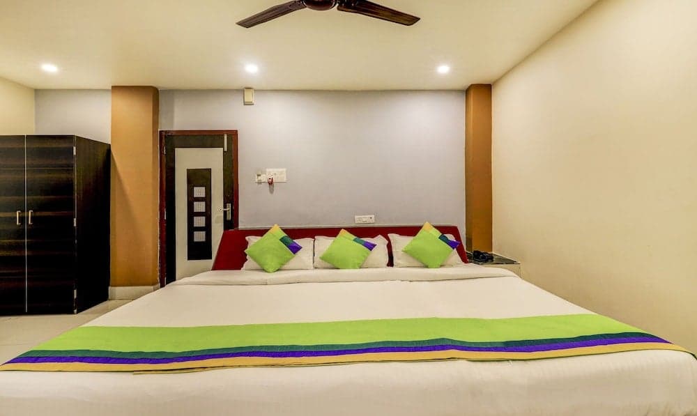 A cozy hotel room features a neatly made bed with vibrant green and purple accents, wooden furnishings, and warm lighting, ideal for travelers seeking comfort and relaxation.