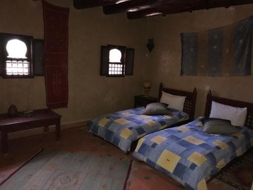 A cozy, rustic bedroom with traditional decor, featuring two beds with blue and yellow patchwork quilts, arched windows with wooden lattices, and earthy tones, ideal for travelers seeking an authentic cultural experience.
