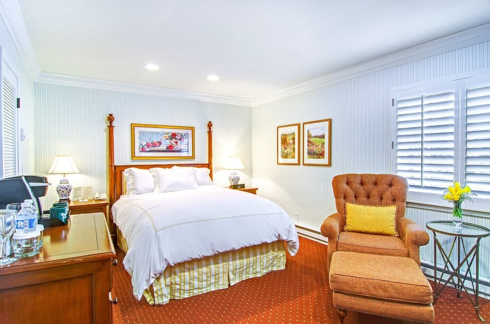 A cozy hotel room features a neatly made bed with white linens, an orange tufted armchair by the window, framed artwork on striped walls, and a warm, inviting atmosphere ideal for travelers seeking comfort and relaxation.
