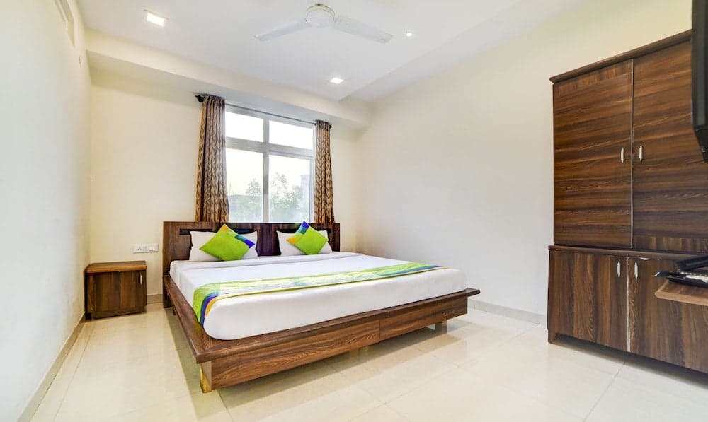 A cozy hotel room features a neatly made bed with colorful pillows, wooden furniture, and a bright window, creating a welcoming atmosphere ideal for travelers seeking comfort and simplicity.