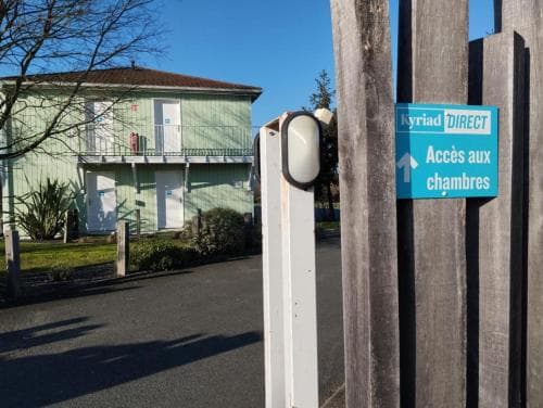 A serene hotel entrance features a Kyriad Direct signpost directing guests to the rooms, set against a backdrop of a quaint two-story building under a clear blue sky, ideal for travelers seeking accommodation details.