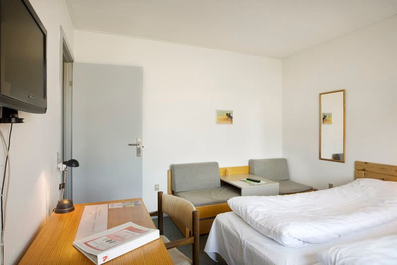 A cozy hotel room features a neatly made bed, wooden desk with books, comfortable seating area, and wall-mounted TV, ideal for travelers seeking a simple yet functional accommodation.
