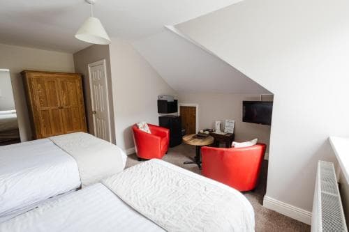 A cozy attic-style hotel room features two neatly made beds, vibrant red armchairs, a small desk area, and a TV, ideal for travelers seeking comfort in a compact space.