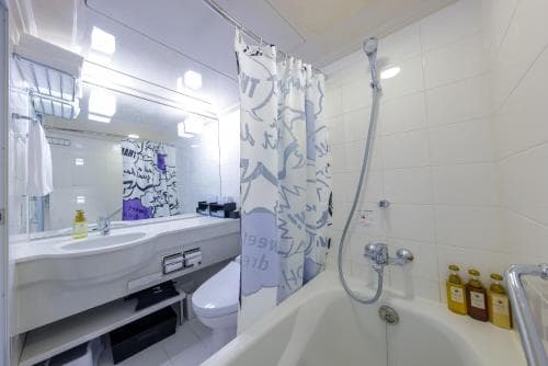 A modern bathroom with white tiles, a bathtub, a shower curtain featuring artistic designs, and toiletries on the side, ideal for travelers seeking clean and stylish accommodations.