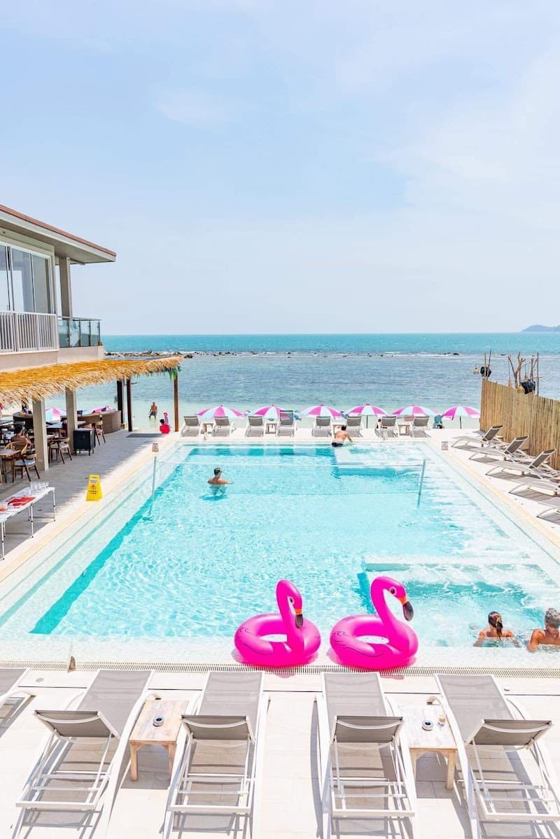 A vibrant beach club scene features a sparkling pool with pink flamingo floats, lounge chairs, and colorful umbrellas overlooking the turquoise sea, perfect for a relaxing tropical getaway.