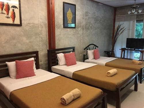 A cozy guesthouse room features neatly arranged twin beds with red cushions, a serene Buddha artwork, and a view of lush greenery outside, ideal for travelers seeking a tranquil and budget-friendly accommodation.