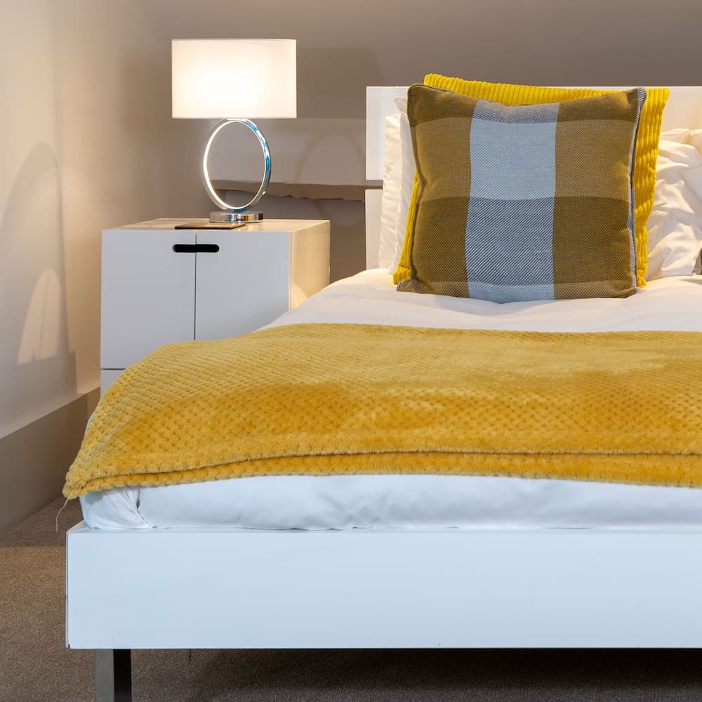 A cozy hotel room features a neatly made bed with a vibrant yellow throw and plaid pillow, complemented by a modern bedside table with a glowing lamp, ideal for travelers seeking comfort and style.