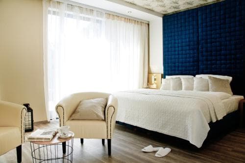 A cozy hotel room features a plush bed with a navy blue headboard, soft beige armchairs, and large windows draped in sheer curtains, creating a serene and inviting atmosphere ideal for relaxation during a getaway.