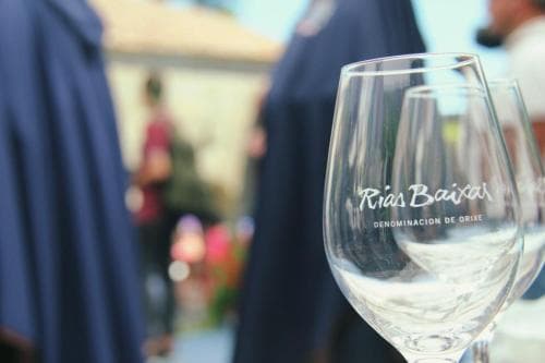 A close-up of a Rías Baixas wine glass in focus against a blurred outdoor setting with people in dark robes, evoking a serene and sophisticated wine-tasting experience ideal for culinary travelers exploring Spanish vineyards.