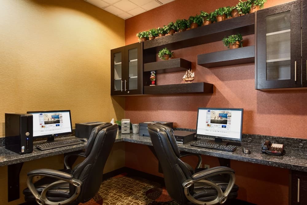 A cozy hotel business center features two ergonomic chairs at granite desks equipped with computers, surrounded by warm-toned walls adorned with shelves of greenery and decorative items, ideal for travelers needing a professional workspace.