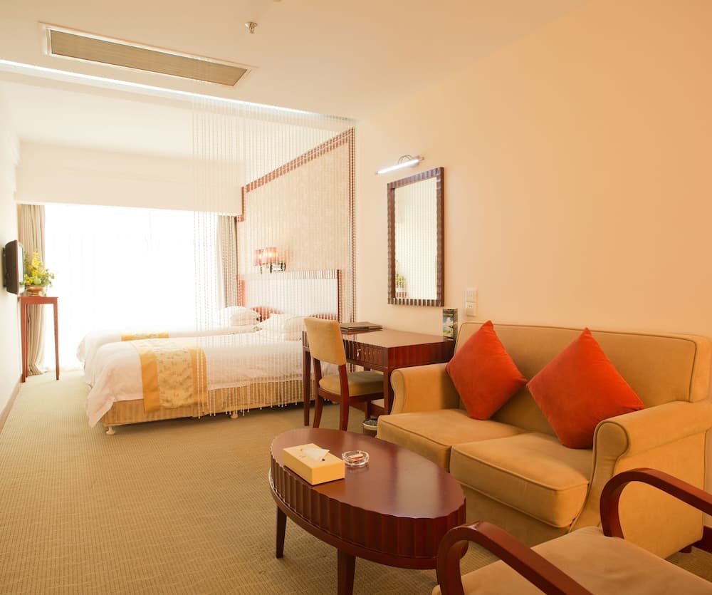 A cozy hotel room features a neatly made bed behind sheer curtains, a comfortable sofa with orange pillows, and a wooden coffee table, creating a warm and inviting atmosphere ideal for travelers seeking relaxation.