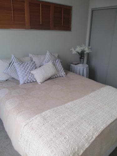 A cozy bedroom with a neatly made bed adorned with patterned pillows, a vase of white flowers on a side table, and wooden shutters, ideal for travelers seeking a serene and comfortable stay.