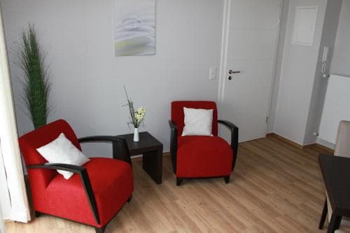 A cozy hotel room features vibrant red armchairs with white cushions, a small table with flowers, wooden flooring, and minimalist decor, ideal for travelers seeking comfort and simplicity.