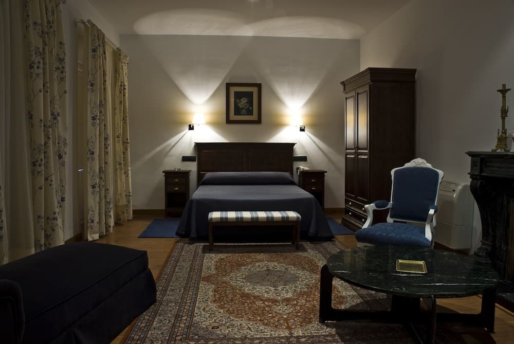 A cozy hotel room features a neatly made bed under warm wall lights, floral curtains, antique furniture, and a patterned rug, creating an inviting atmosphere ideal for travelers seeking comfort and charm.